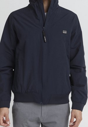 Homme portant une veste zippée bleu marine avec capuche et un petit patch carré sur la poitrine gauche, associée à un pantalon gris clair.