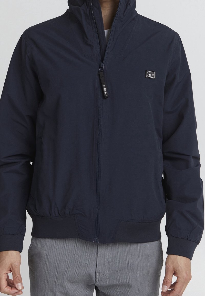 Homme portant une veste zippée bleu marine avec capuche et un petit patch carré sur la poitrine gauche, associée à un pantalon gris clair.