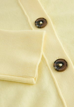 Prim-plan al unei țesături de tricot de culoare galben-palid, cu manseta pliată și două butoane maro marmorat coase pe un articol de îmbrăcăminte.