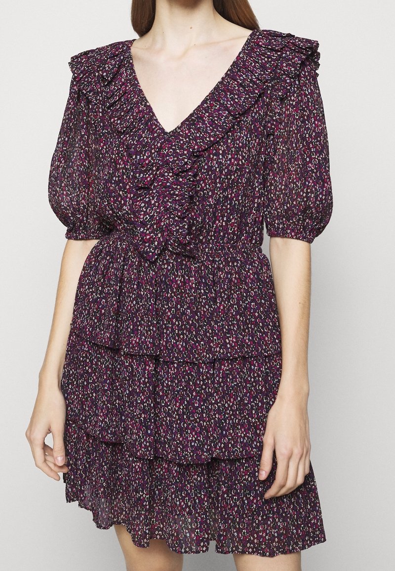 Femme portant une robe fleurie violet foncé avec un décolleté en V à volants, des manches ballon et une jupe à plusieurs couches, debout devant un fond uni.