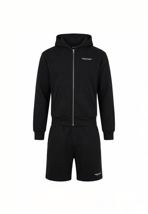 Młody mężczyzna stoi twarzą do przodu, ubrany w czarną bluzę z kapturem na suwak, czarne szorty i białe tenisówki na białym tle.