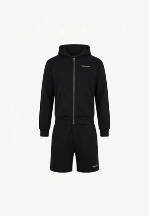 Młody mężczyzna stoi twarzą do przodu, ubrany w czarną bluzę z kapturem na suwak, czarne szorty i białe tenisówki na białym tle.