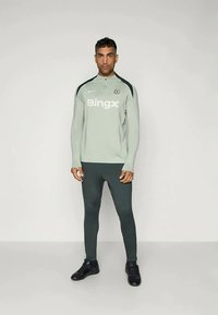 Lys grå treningsoverdel med svarte detaljer og logo, kombinert med mørkegrønne tettsittende leggings og svarte treningessko. Passer for sportstøy.