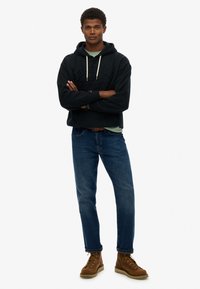 Sudadera de algodón negra con cordones, jeans de mezclilla y botas de cordones marrones. El modelo está de pie con los brazos cruzados, vistiendo una camiseta verde claro debajo.