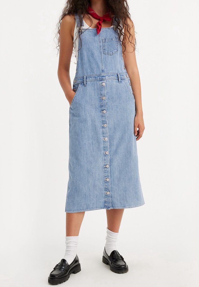 Femme portant une robe midi en denim bleu clair avec des boutons sur le devant, des poches, des chaussettes blanches, des mocassins noirs et un foulard rouge autour du cou.