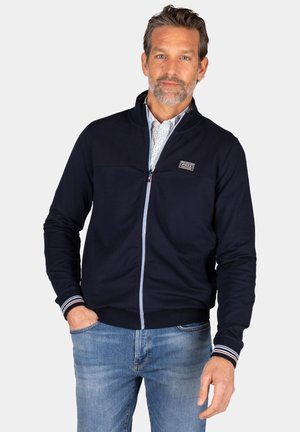 Marine blauwe ritsjack, gemaakt van gladde stof, met een staande kraag en gestreepte boorden. Een logo-applicatie is zichtbaar op het borstpaneel.