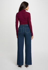 Burgundijska rebrasta turtleneck pulover kombiniran s trapericama širokih nogavica tamnoplave boje s visokim strukom, koje imaju dva stražnja džepa i srebrne detalje.
