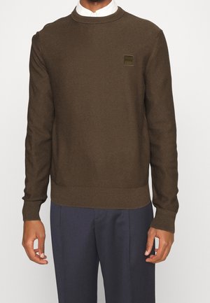 Pull en tricot marron avec un motif texturé, col rond et poignets côtelés. Présente un petit patch logo vert sur la poitrine.