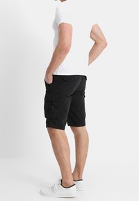 Shorts cargo noirs en tissu léger avec plusieurs poches latérales et une coupe décontractée. Portés avec un t-shirt blanc ajusté et des baskets.