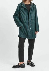 Mörkgrön vattentät jacka med huva, tryckknappsstängning, två framfickor och långa ärmar, kombinerad med svarta jeans och loafers.