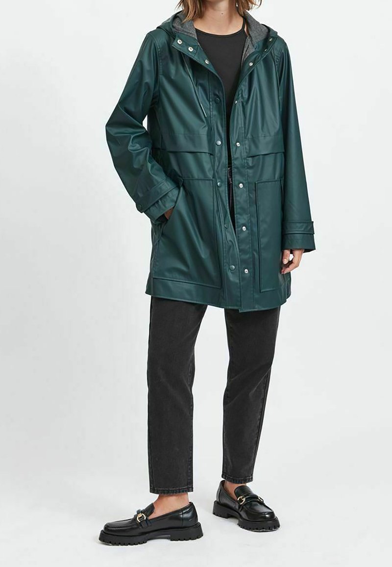 Mörkgrön vattentät jacka med huva, tryckknappsstängning, två framfickor och långa ärmar, kombinerad med svarta jeans och loafers.