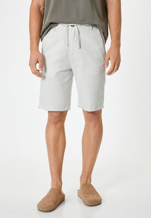 Pantalones cortos gris claro de tela texturizada con cintura ajustable mediante cordón, bolsillos laterales, dobladillo enrollado y combinados con zapatos marrones de tipo slip-on.