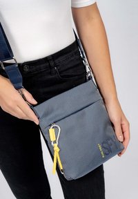 Borsa a tracolla blu con texture, dotata di una tasca frontale con cerniera, accentuata da un dettagli giallo sul tirante della cerniera e una tracolla regolabile, tenuta in mano da una persona.