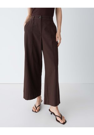 Frau trägt dunkelbraune, hoch taillierte, weit geschnittene Hose mit vorderen Falten und schwarze Sandalen mit kleinen Absatz und Zehensteg, steht mit den Händen in den Taschen.