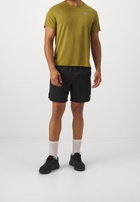 Camiseta de manga corta verde oliva, pantalones cortos deportivos negros, calcetines blancos de caña corta, zapatillas de correr negras, con un pequeño logotipo gris en la camiseta y los pantalones cortos.