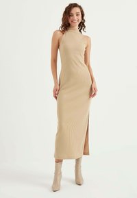 Abito midi in maglia beige con scollo alto e spacco laterale, abbinato a stivaletti con tacco alti fino alla caviglia in tonalità chiara.