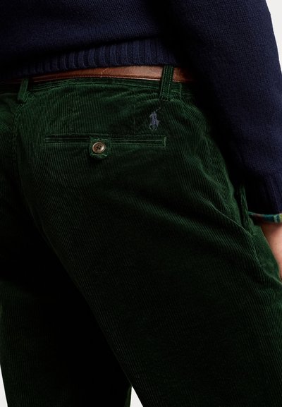 Gröna kordbyxor med en texturerad yta, försedd med en bakficka med knapp och en liten broderad logotyp. En brun bälte är synlig.
