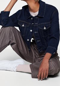 Mörkblå denimjacka i cropped-stil med silverfärgad hårdvara och två frampocket, kombinerad med bruna rutig byxor och vita sneakers.