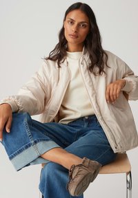 Femme assise sur une chaise, jambes croisées, portant une veste bomber beige, un pull crème, un jean bleu retroussé et des chaussures en suède marron.