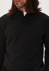 Pull-over en tricot noir avec col, présentant une patte de boutonnage à trois boutons et un tissu lisse et texturé. Manches longues, coupe classique.
