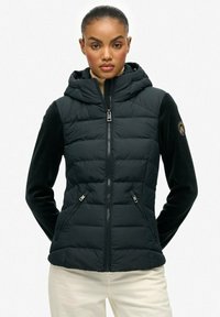 Chaqueta acolchada negra con capucha, que presenta un acabado de textura acolchada, bolsillos laterales con cremalleras y mangas de tela lisa en contraste.