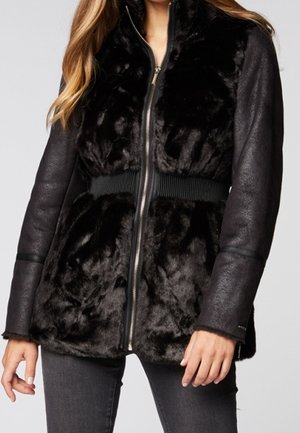 Mujer con una chaqueta negra con cremallera, con un panel frontal peludo y mangas largas de textura suave, combinada con vaqueros oscuros.