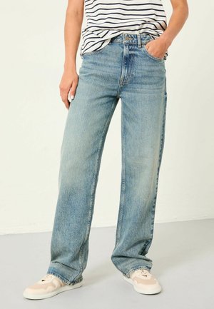 Person iført lyseblå vidde jeans, hvide og beige sneakers og en hvid og navystribet skjorte med den ene hånd i lommen.