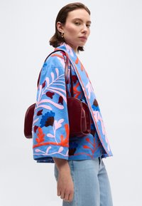Γυναίκα ντυμένη με μπλε μπουφάν με λουλουδάτο μοτίβο και ροζ και πορτοκαλί λεπτομέρειες, ανοιχτόχρωμα τζιν και κρατώντας μια σκούρα κόκκινη τσάντα ώμου.