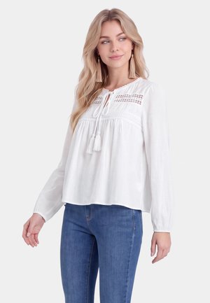Young woman wearing a white long-sleeve blouse with lace details and tassel ties, paired with blue jeans, looking to the side.