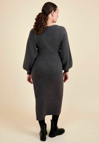 Grau gestricktes Kleid mit langen Ballonärmeln, tailliertem Schnitt und Knielänge. Getragen mit schwarzen Stiefeletten, glatte Textur, subtile Details.