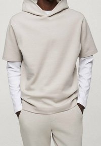 Man wearing beige short-sleeve hooded top over white long-sleeve shirt and matching beige pants with hands in pockets.