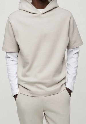 Uomo che indossa una maglietta a maniche corte con cappuccio beige sopra una camicia a maniche lunghe bianca e pantaloni beige coordinati, con le mani nelle tasche.