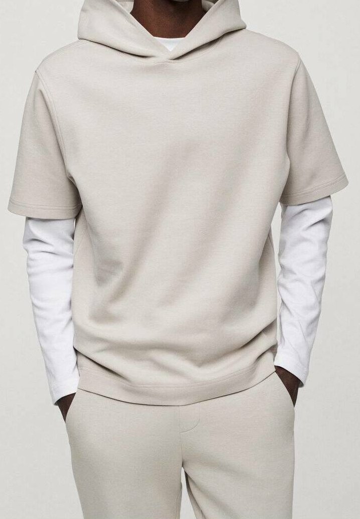 Man wearing beige short-sleeve hooded top over white long-sleeve shirt and matching beige pants with hands in pockets.