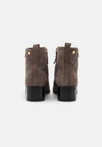 Botines de ante marrón con punta redondeada, tacón bajo de bloque y detalles de cremallera dorada, mostrados desde atrás sobre un fondo blanco.