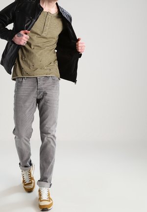 Chaqueta de cuero negra, camiseta henley verde oliva desgastada, vaqueros grises y zapatillas marrones y blancas con una superficie texturizada.