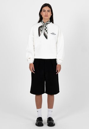 Kvinde iført hvid sweatshirt med "Prohibited"-logo, ternet halstørklæde, sorte shorts til knæet, hvide sokker og sorte loafers, stående stille.