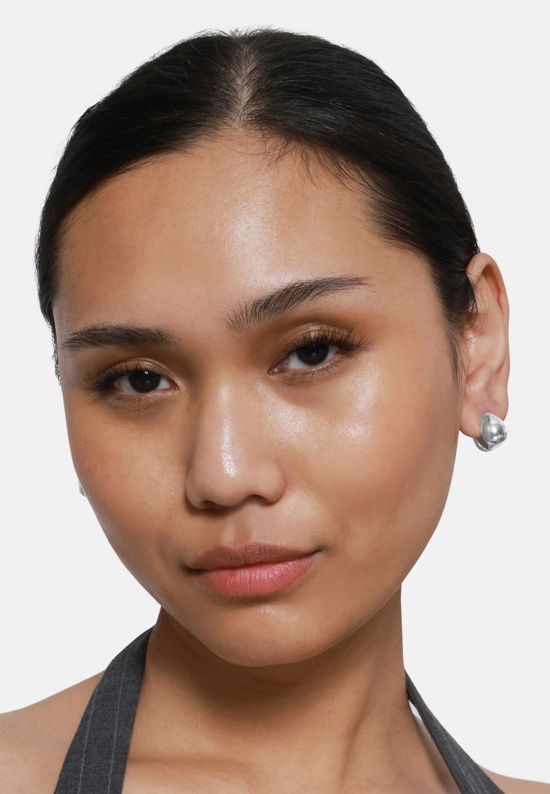 Orecchini in metallo argentato con una forma organica, alta lucentezza e superficie liscia, indossati da una persona con capelli scuri e labbra neutre su uno sfondo bianco.