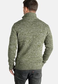 Suéter de punto verde con un cuello alto, que presenta un patrón texturizado y moteado. Puños y dobladillo acanalados. Combinado con jeans gris oscuro.