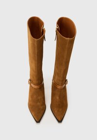 Bottes en suede marron jusqu'au genou avec des pointes relevées, accentuées par une sangle marron et une boucle dorée, avec une doublure intérieure lisse.