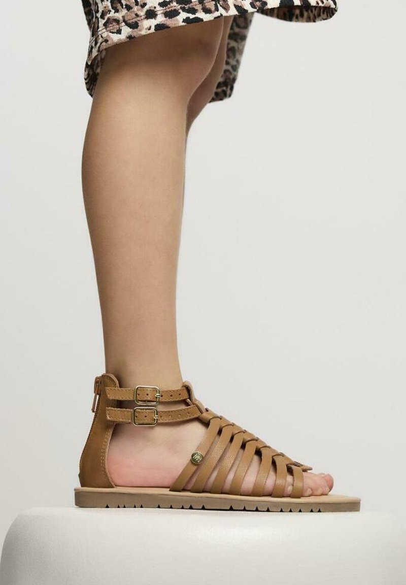 Sandalia de cuero marrón con tiras, dos hebillas ajustables, suela plana y diseño de punta abierta. Las tiras texturizadas crean un patrón trenzado.