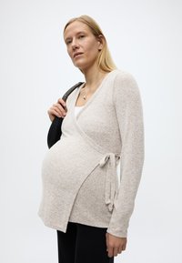 Έγκυος γυναίκα με μακριά ξανθά μαλλιά, φορώντας μπεζ πουλόβερ τύπου wrap και κρατώντας μια μαύρη τσάντα ώμου σε λευκό φόντο.