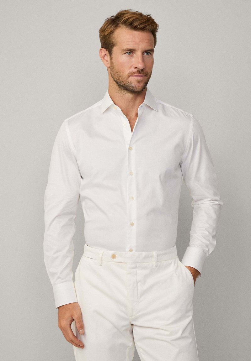 Camicia bianca a maniche lunghe, realizzata in tessuto morbido con colletto classico e vestibilità su misura, abbinata a pantaloni bianchi.