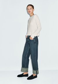 Maglione grigio chiaro, texture; jeans oversize blu scuro con orli risvoltati; scarpe slip-on nere con una finitura testurizzata. Design semplice e aderente.