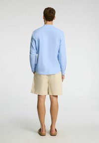 Svetlo modra srajca z dolgimi rokavi z diskretno teksturo, v kombinaciji s bež kratkimi hlačami. Model prikazan s hrbtne strani, obut v rjave sandale.