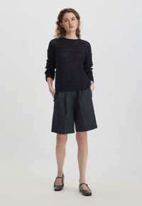 Navy gestrickter Pullover mit offener Maschenstruktur, Rundhalsausschnitt und langen Ärmeln, kombiniert mit dunkelgrünen maßgeschneiderten Shorts und schwarzen Mary Jane Schuhen.