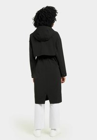 Veste imperméable noire avec un crop top, capuche, taille cintrée et dos long. Associée à un pantalon blanc large et des baskets blanches.