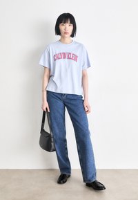 Hellblaues Baumwoll-T-Shirt mit rotem "CALVIN KLEIN"-Schriftzug, kombiniert mit blauen Jeans im Straight-Leg-Schnitt. Das Model hält eine schwarze Schultertasche.