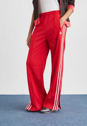 Personne portant un pantalon de survêtement Adidas rouge à bandes blanches, une main dans la poche, associé à des chaussures argentées et une veste noire et rouge.