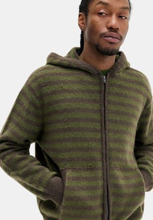 Homme portant un sweat à capuche à rayures vertes et marron avec fermeture éclair, les mains dans les poches, regardant sur le côté sur fond uni.
