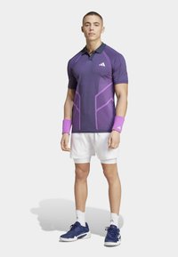 Polo deportivo morado con cuello negro, que presenta patrones geométricos morados; combinado con pantalones cortos blancos y zapatillas deportivas azules. Incluye muñequeras.